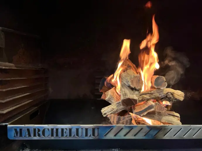 A heavy-duty mild steel Marchelulu braai insert with a bright wood fire, demonstrating a custom fit inside a brick braai.