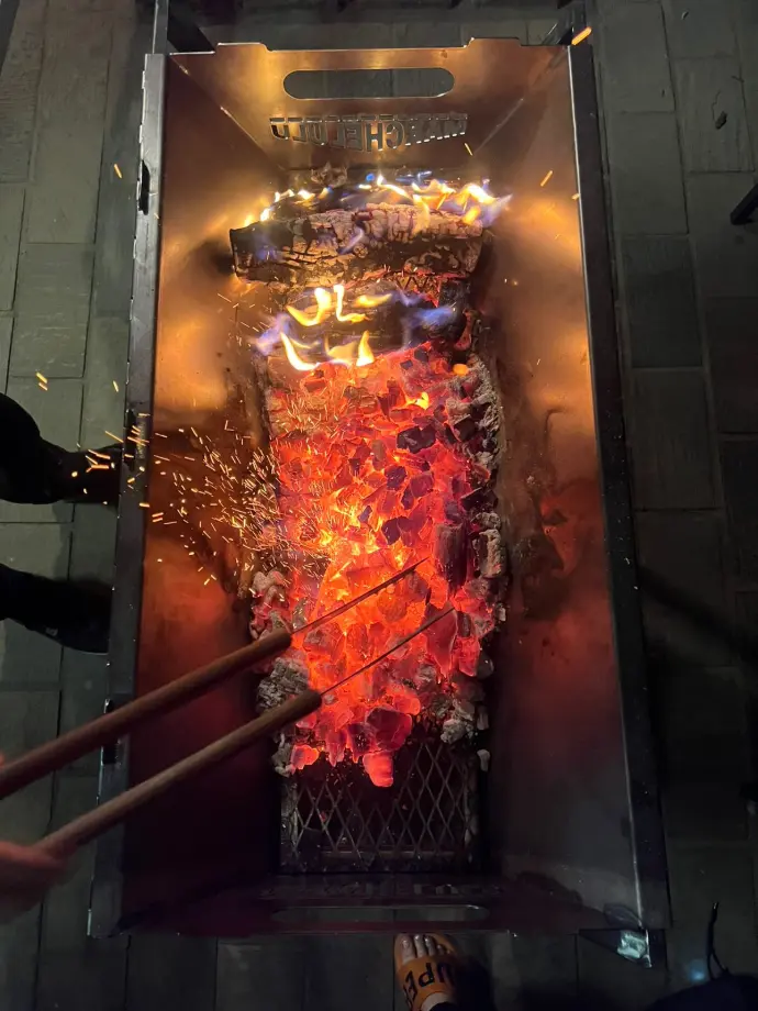 Top-down view of a Marchelulu mild steel V-Bucket with glowing hot coals, showing the heavy-duty coal grid and sparks from the fire.