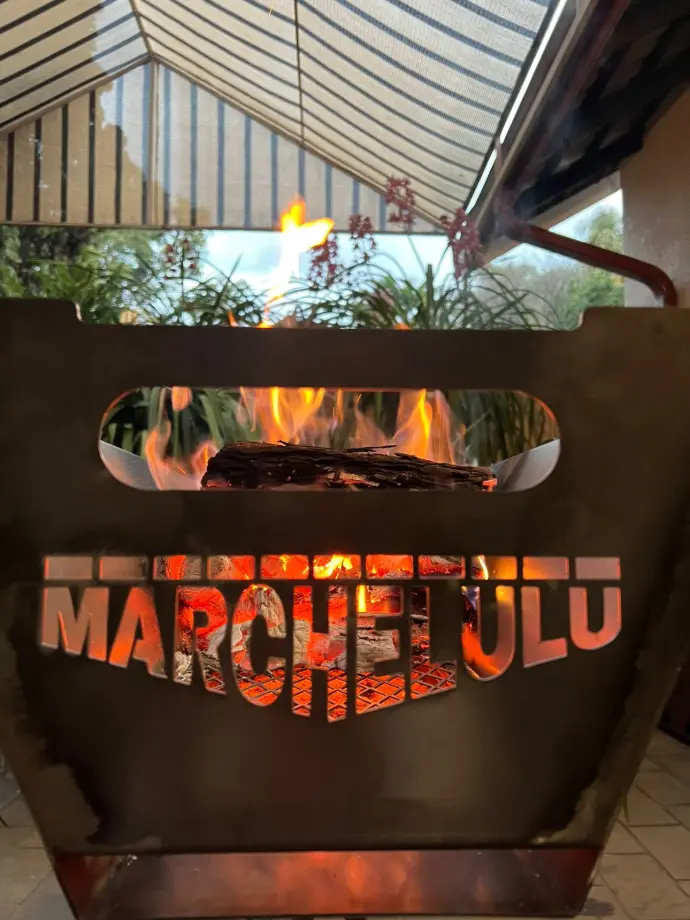 Close-up of the laser-cut Marchelulu logo on a braai bucket, with bright flames and glowing coals visible through the lettering.