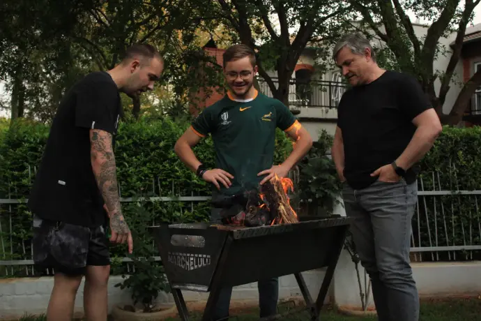 The Marchelulu co-founders enjoying a classic South African braai with a friend wearing a Springbok jersey, gathered around the V-Bucket on its freestanding stand in a garden.